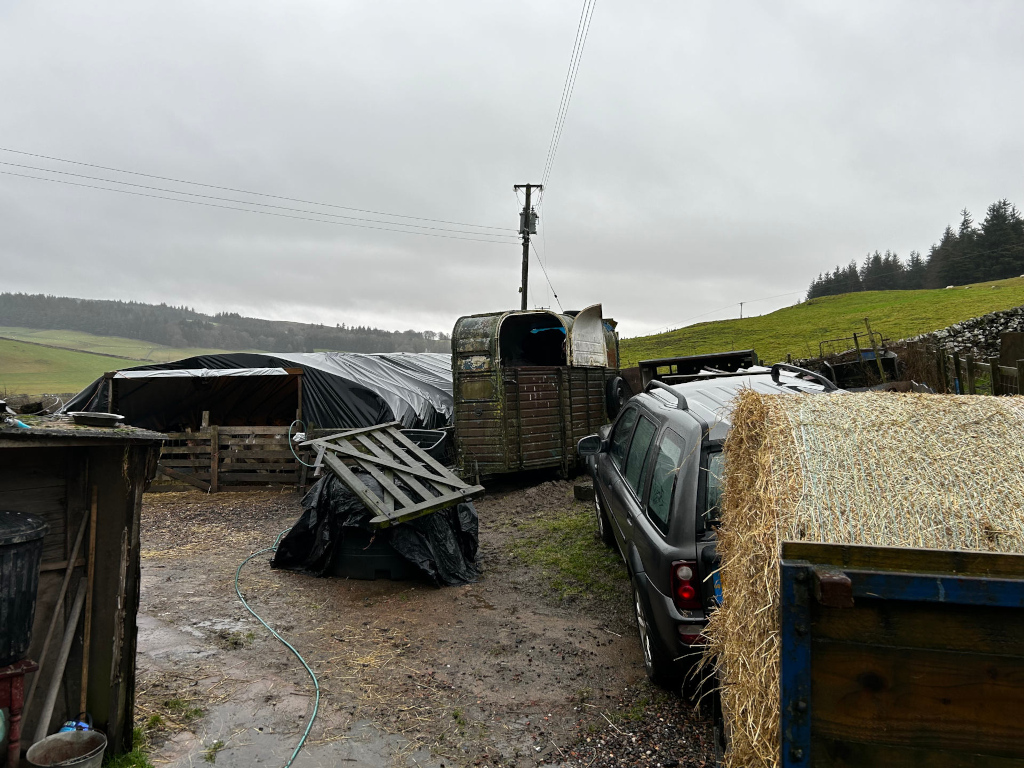 Farmyard scene on an overcast day. A dark-coloured Land Rover is parked partially obscured by a large round bale of hay. In the background, there is a livestock trailer, a structure covered with a tarp, and various farm implements.