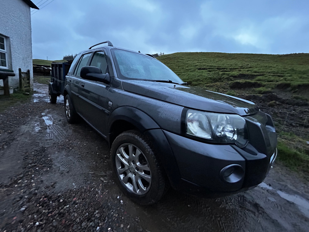 Dark grey Land Rover Freelander 1 parked on a muddy gravel driveway outside a stone building. The Land Rover is towing a small trailer. The background shows a grassy hillside under a cloudy sky. The overall mood is somewhat sombre due to the overcast lighting.