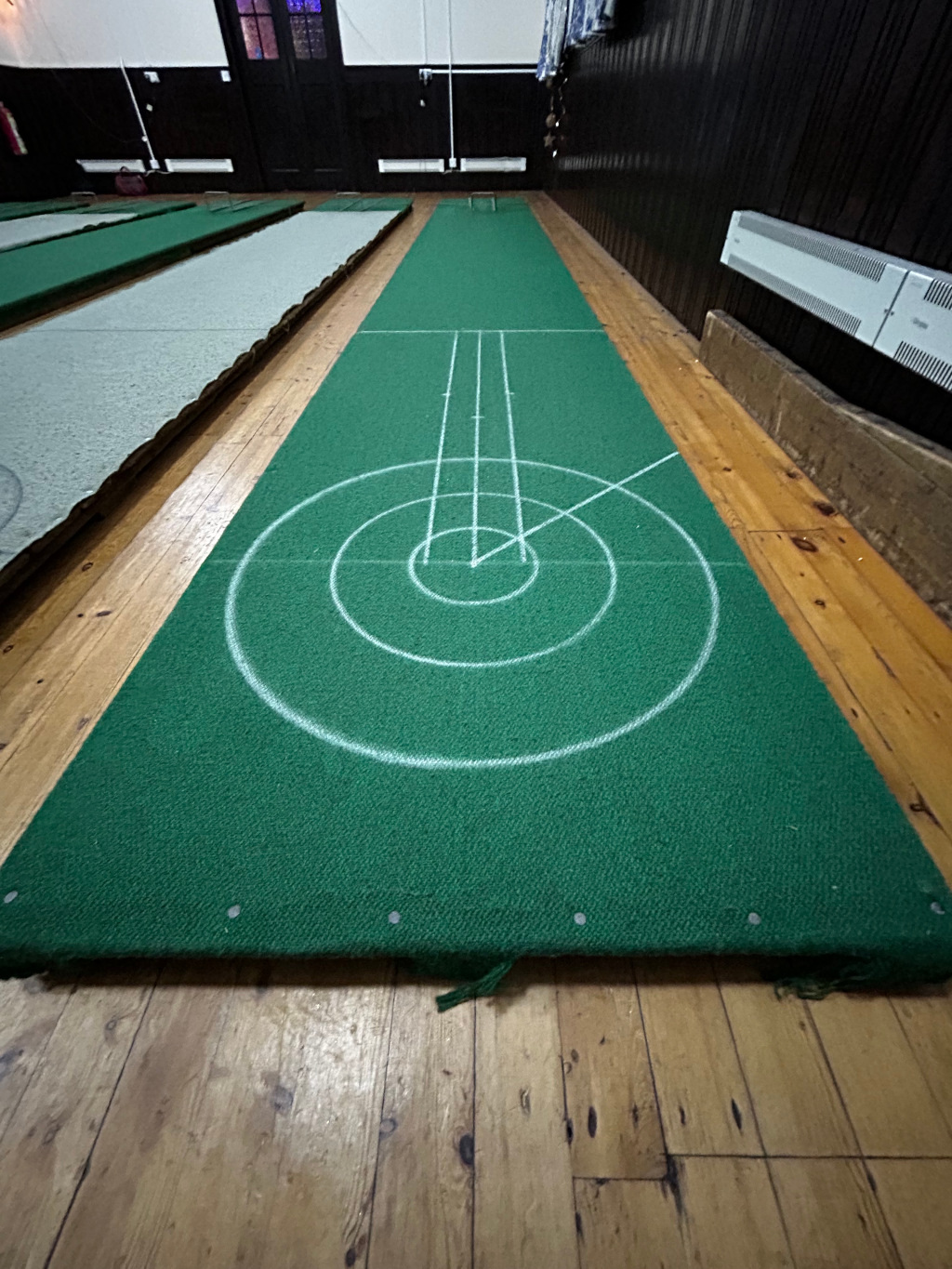 Green carpet with white lines forming concentric circles and lines, typical of a bowls rink or a similar game. The carpet is laid on a wooden floor, and other similar mats are visible in the background. The overall impression is of an indoor game space.