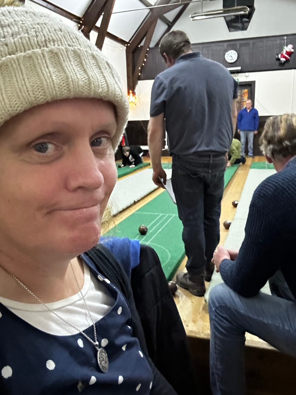 Selfie taken by a Leonie wearing a beige knitted hat and a navy blue polka dot dress. She is in a large hall with wooden floors and beams, where a bowls game is taking place. Several people are visible in the background, some playing bowls, and others observing. Leonie is in the foreground appearing to be slightly amused or smirking, perhaps observing the game. The overall mood is casual and relaxed, possibly suggesting a social event.