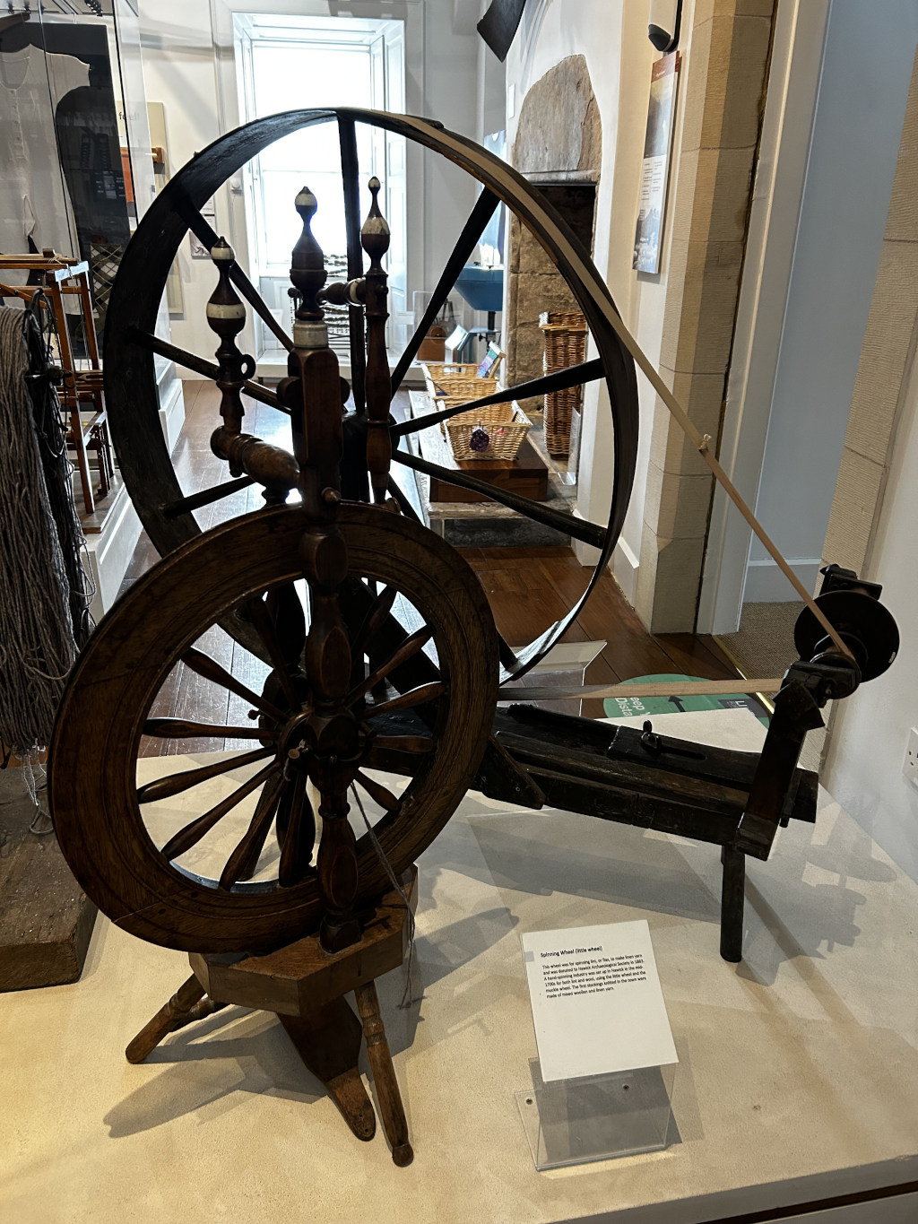 Antique spinning wheel, a device used for spinning yarn or thread from natural fibres. The wheel is made of dark wood, with a large outer wheel and a smaller driving wheel. It's displayed in what appears to be a museum setting, with a small information plaque describing its history and function. The background shows a room with other historical artefacts visible.