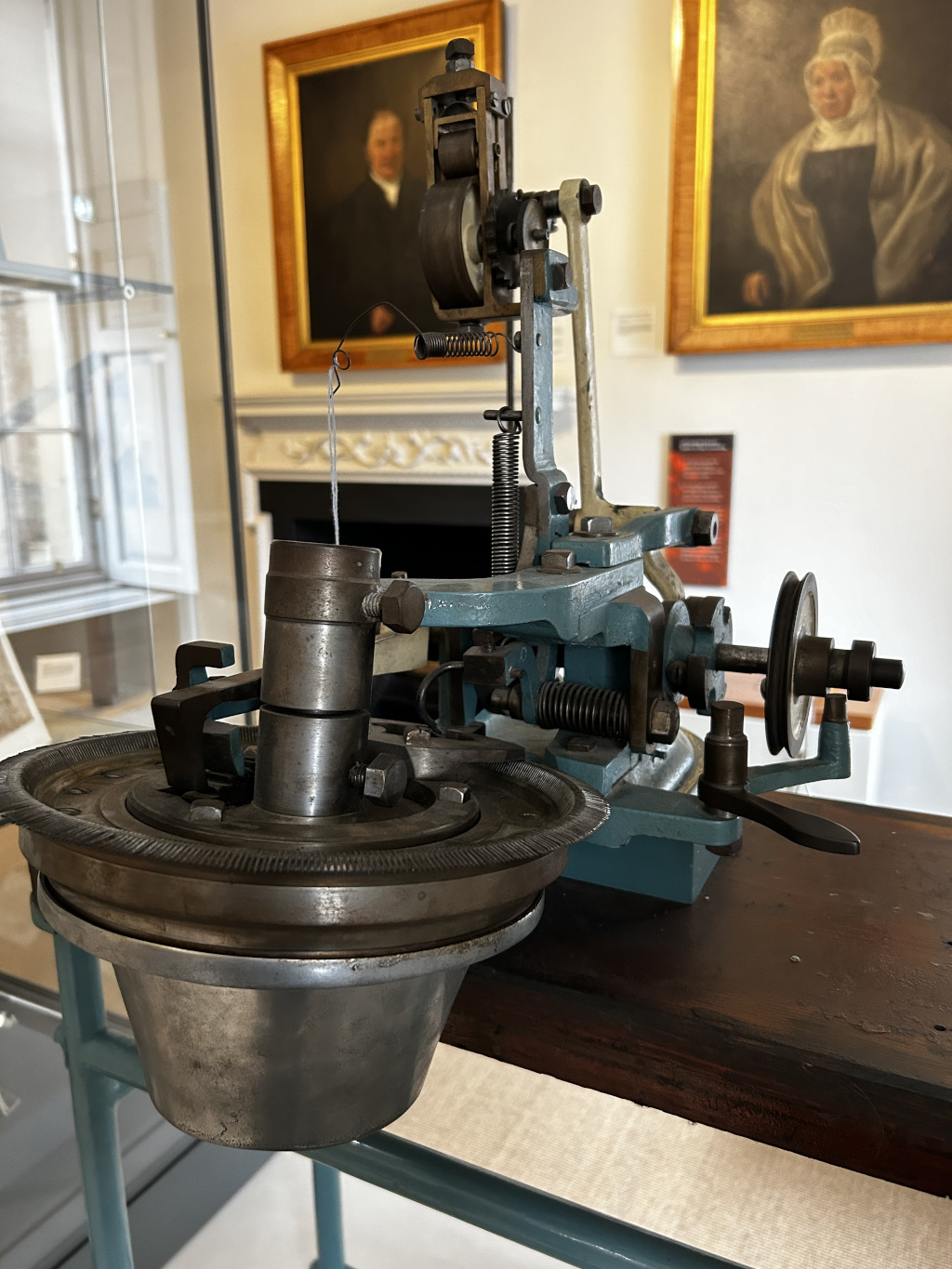 Close-up view of an antique textile machine, possibly a type of yarn-testing or winding device. The machine is teal-coloured metal with various gears, wheels, and levers visible. A thin thread is partially visible, suggesting the machine's function. The machine sits on a dark wooden table, and in the background, there are two framed portraits of people in period clothing, suggesting a historical setting, possibly a museum or historical building.