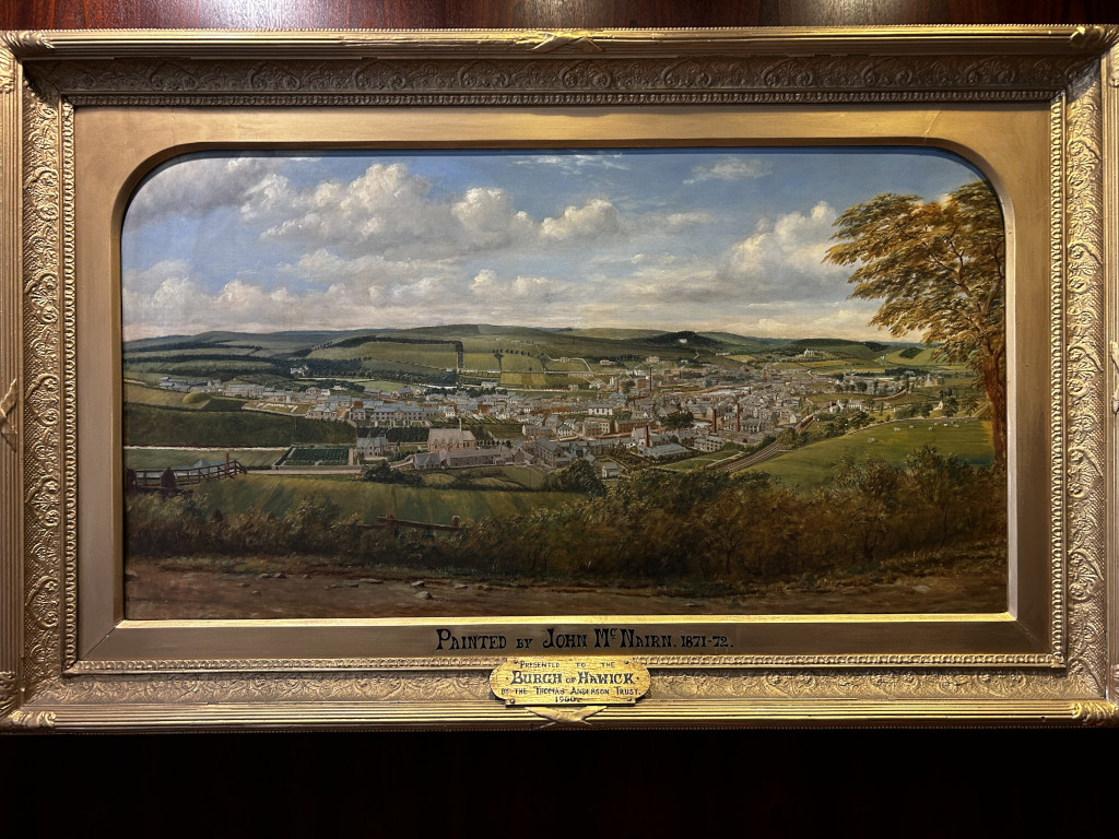 Oil painting on canvas, in a gilded frame, depicting a panoramic view of Hawick, Scotland, as seen from an elevated vantage point. The painting showcases the town's layout, architecture, and surrounding landscape, including fields and rolling hills. The style is realistic and detailed, capturing the atmosphere of a 19th-century town. The painting is dated 1871-72 and attributed to artist John Mc Nairn. The inscription indicates it was a gift to the town of Hawick.