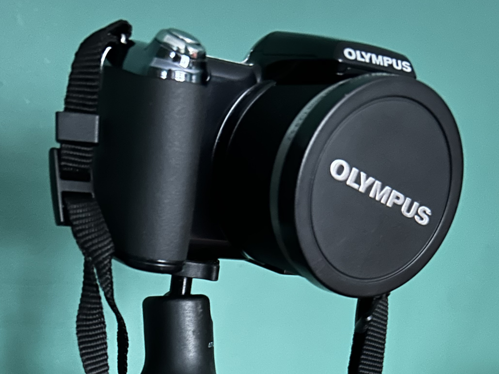 Black Olympus digital camera mounted on a small tripod. The camera lens cap is on. The background is a teal-coloured wall.
