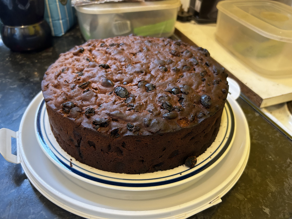 Round, dark brown fruitcake, generously studded with dark raisins or currants. The cake is presented on a white cake stand with a blue trim, which sits on a larger white plastic serving base. The background is blurry but shows a kitchen counter with various containers and appliances. The overall impression is one of a freshly baked, homemade fruitcake, possibly ready for serving.