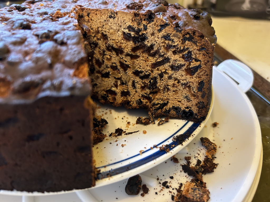 Partially eaten fruitcake on a white plate. A large slice has been cut, revealing the dense, dark fruit-filled interior. Crumbs are scattered on the plate and around the base of the cake.