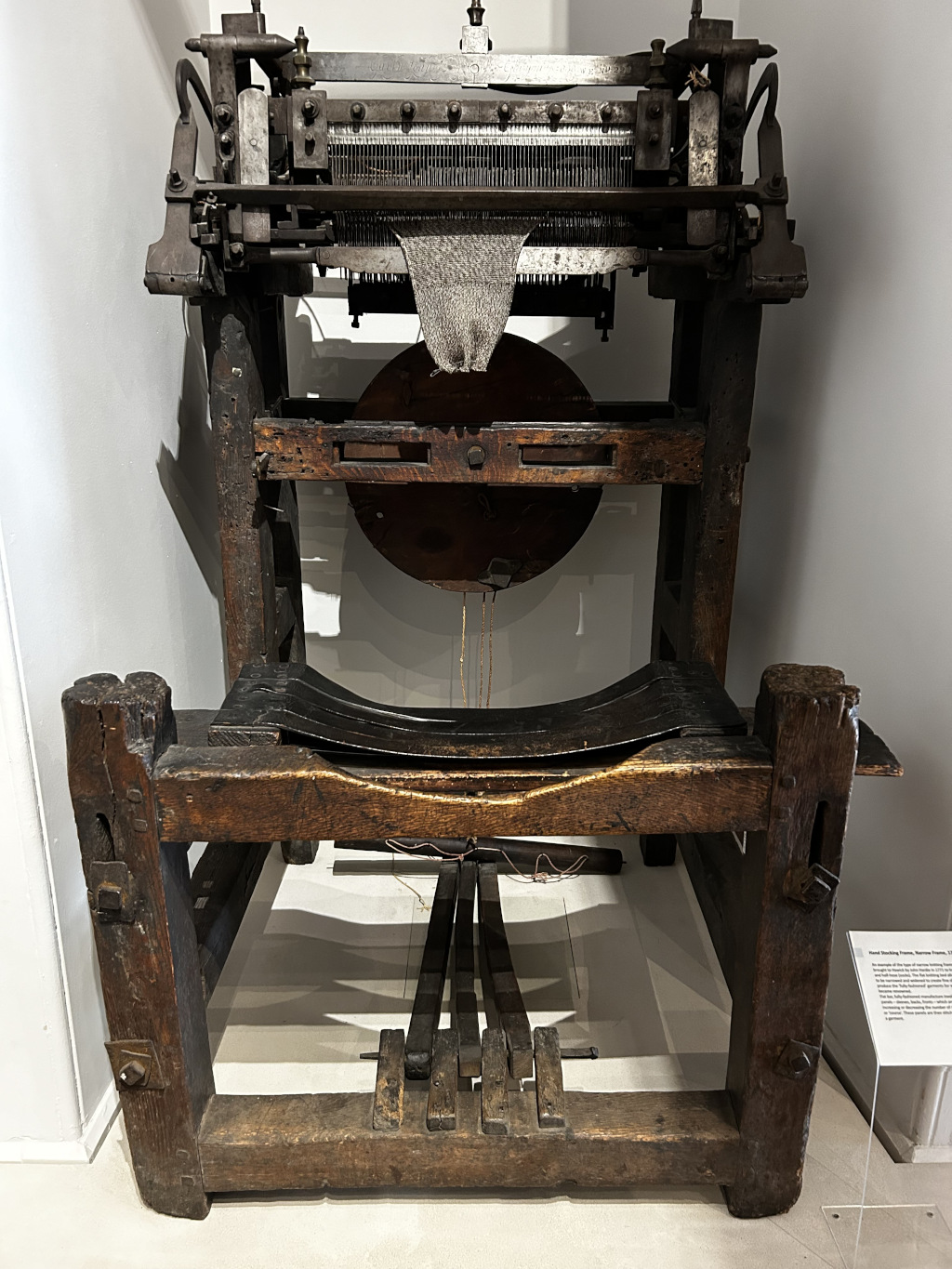 Historical, wooden stocking frame, a machine used for knitting stockings. The frame is made of dark wood, showing signs of age and wear. A partially knitted piece of fabric is visible within the machine's mechanism. The overall impression is one of age and industrial history.