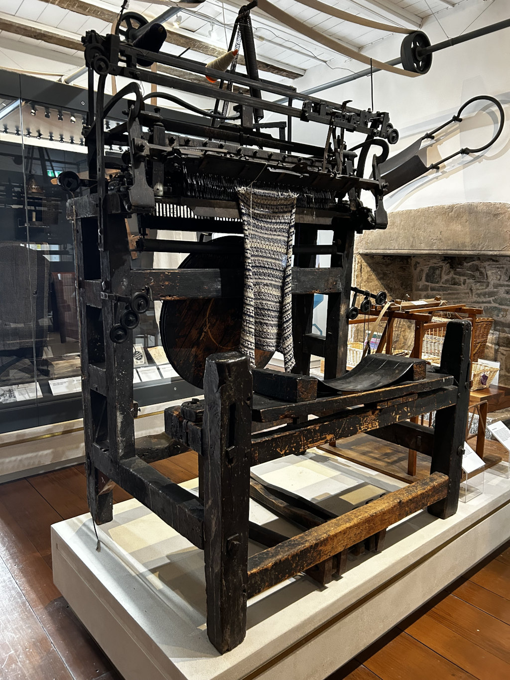 Large, old, wooden loom displayed in a museum setting. The loom appears to be antique, showing significant wear and signs of age. A partially woven piece of fabric is visible on the loom. The surrounding environment suggests a historical exhibit focused on textile production.