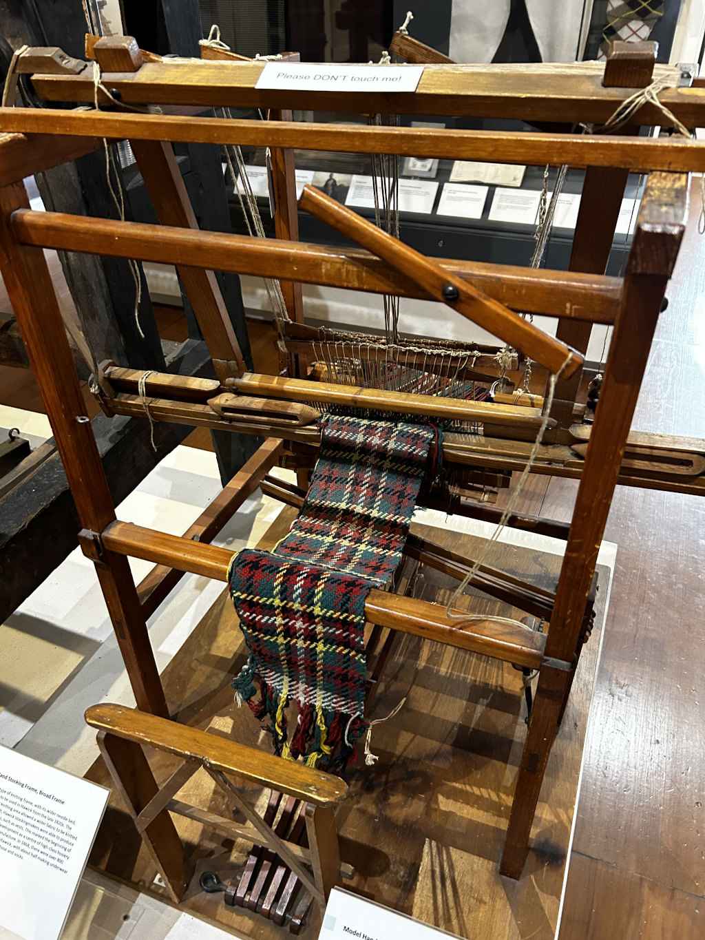 Small wooden model of a hand loom, a device used for weaving textiles. A partially completed piece of plaid fabric is visible on the loom. A sign indicates that the loom is not to be touched. The setting appears to be a museum display, with informational text cards nearby describing the loom and its historical context. The overall impression is one of historical preservation and the display of craftsmanship.