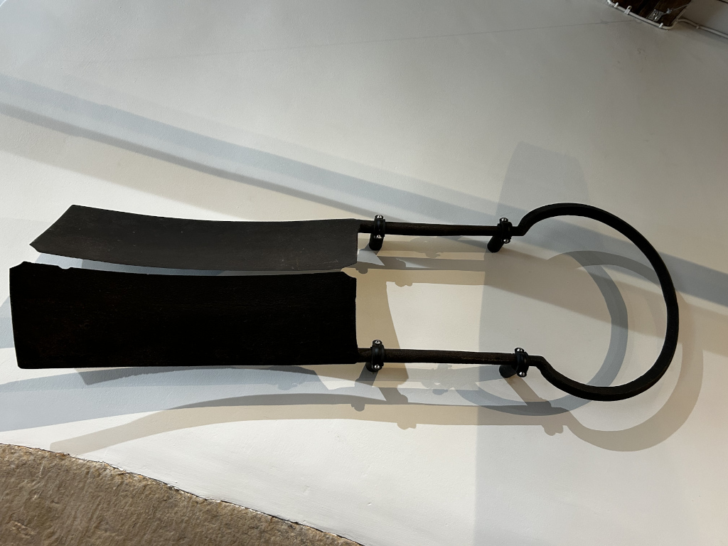 Fragmented, dark-coloured metal object, possibly a piece of machinery or a tool, mounted on a light-coloured wall. The object consists of two flat, blade-like pieces connected by a curved metal framework. One of the blade-like pieces appears slightly damaged or broken. The overall impression is one of age and possible incompleteness.