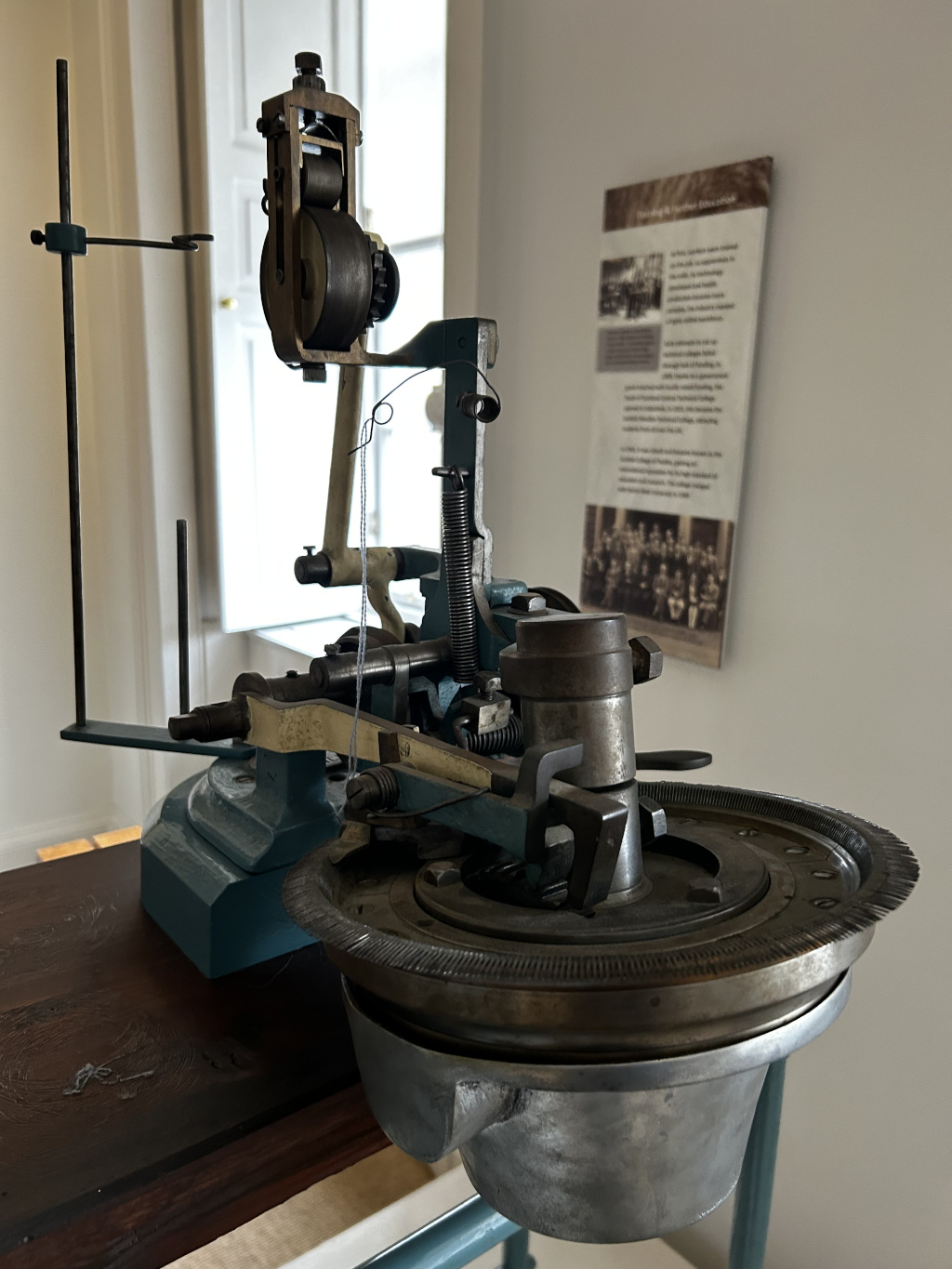 Close-up view of an antique, teal-coloured machine, possibly used in textile manufacturing or a related process. The machine has various metal components, gears, and a circular base. A thin white thread is partially visible, suggesting it is a device that uses thread in its operation. The machine sits on a dark brown wooden table. In the background, a wall-mounted informational panel with historical photos is partially visible.
