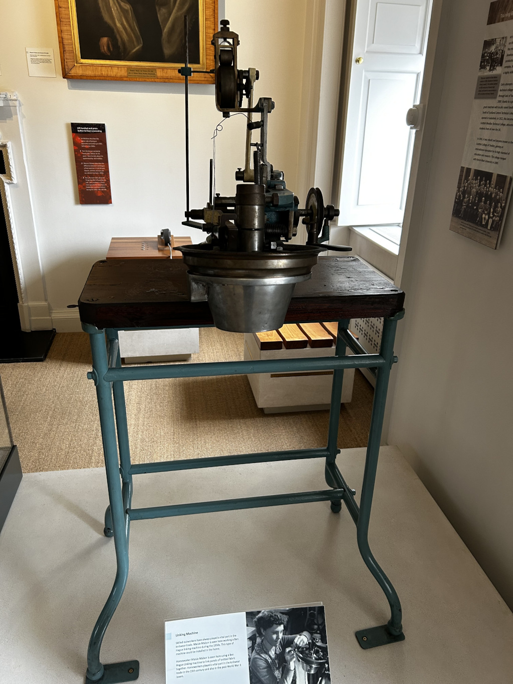 Vintage Linking Machine displayed on a teal-coloured metal stand in what appears to be a museum setting. The machine itself is dark metal and seems to be a specialised piece of equipment used for some kind of crafting or manufacturing process, possibly involving thread or wire judging by the visible spool and wire guide. There is an informational card next to the machine with a brief description and a historical photograph of a woman operating a similar device. The background includes walls with framed artwork and informational displays, typical of a museum exhibit. The overall tone is one of historical preservation and industrial heritage.