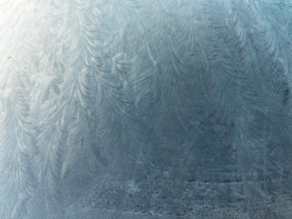 Close-up view of frost patterns on a cold surface, possibly a window or a similar material. The frost forms delicate, feathery designs in shades of light and dark blue-grey, creating a textured and somewhat abstract aesthetic. The overall impression is one of cold, stillness, and the beauty found in natural, ephemeral formations.