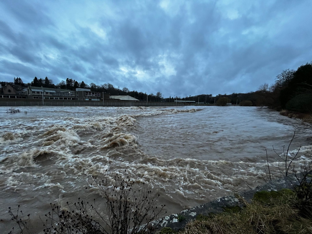 Wide river in flood. The water is brown and turbulent, with whitecaps indicating a strong current. A low stone wall runs along the bottom right of the image, separating the viewer from the river. In the background, buildings and trees are visible, indicating a residential or urban area close to the river. The sky is overcast, adding to the sombre mood of the scene. The overall impression is one of a powerful and potentially destructive natural event.
