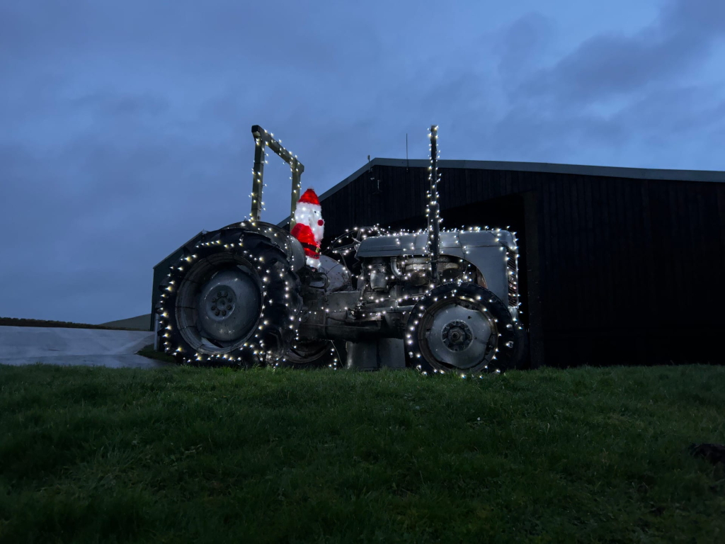Vintage tractor decorated with Christmas lights. A Santa Claus figure is positioned on the tractor's seat. The tractor is parked in front of a dark barn, on a grassy area, at dusk or night. The overall scene evokes a festive, rural Christmas atmosphere.