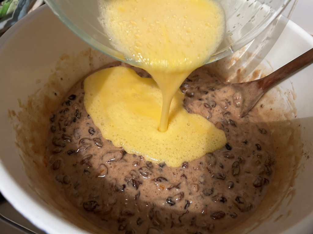 Close-up view of a white bowl containing a mixture of dried fruits and spices. A light yellow liquid, likely a beaten egg mixture, is being poured from a clear glass bowl into the bowl of fruit and spice mixture. A wooden spoon is visible in the bowl. The scene depicts a step in the preparation of a fruitcake or similar baked good.