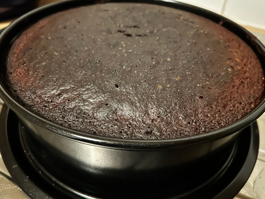 Dark fruit cake, freshly baked and still in its black spring form pan. The cake's surface is slightly domed and has a textured appearance, showing small air bubbles from baking.