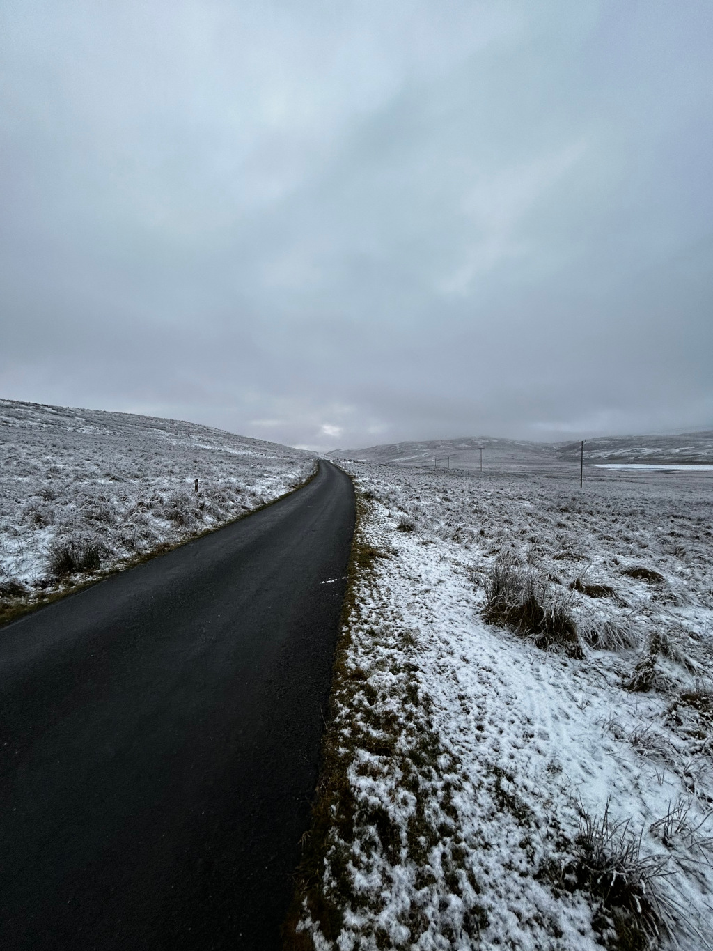 Dark asphalt road winding through a snow-covered landscape under a cloudy sky. The road appears to lead towards a distant, slightly elevated horizon. The overall colour palette is muted, with greys and whites dominating the scene. The snow is patchy, not covering the ground completely. There's a sense of vastness and quietude.