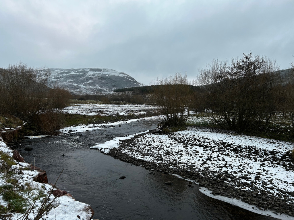 Tranquil river scene in a snowy landscape. A dark, meandering river flows through a flat, snow-dusted valley. The riverbanks are composed of dark rocks and pebbles, sparsely covered with a light layer of snow. Bare, winter trees line the river, and a snow-covered hill rises in the background under a grey, overcast sky. The overall mood is peaceful and somewhat sombre, reflective of a cold, winter day.