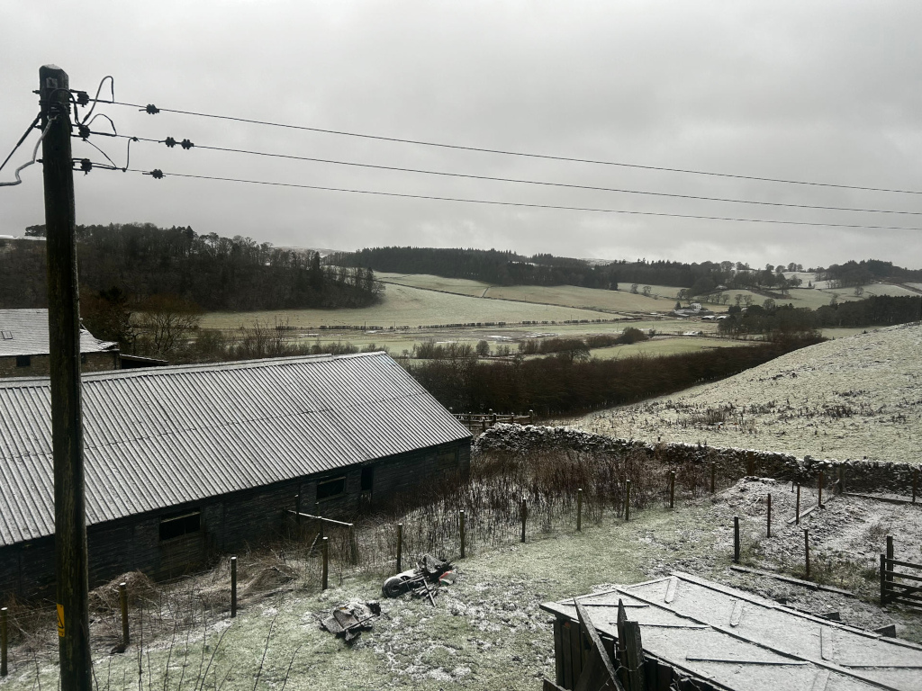 Snow-dusted rural landscape, viewed from a slightly elevated perspective. In the foreground is a long, dark barn with a corrugated metal roof, partially covered in snow. To the right, there are sections of low stone walls, and a partially visible wooden structure, also dusted with snow. A utility pole with power lines is prominent on the left. Beyond the immediate foreground sits a gently rolling landscape of fields and woodlands, all lightly covered in snow under a grey, overcast sky. The overall mood is quiet and somewhat bleak, suggestive of a cold, winter's day in a rural area.