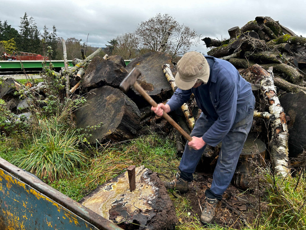 Charlie splitting a large log with a maul and wedge. He is wearing work clothes and a cap. The log is resting on a stump, surrounded by a pile of other logs and wood debris. The setting appears to be outdoors, possibly in a wooded area or lumber yard. The overall impression is one of manual labour and possibly wood preparation.