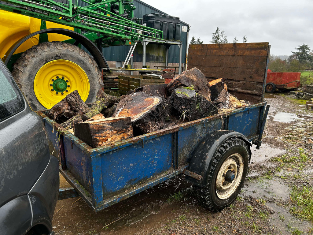 Blue trailer loaded with a pile of chopped wood logs. The trailer is attached to a dark-coloured vehicle, and a portion of a large green agricultural machine is visible in the background. The scene appears to be set in a rural or agricultural setting, possibly a farm or yard, on a wet, muddy day.