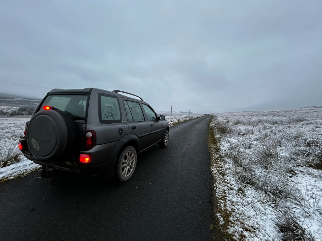 Dark gray Land Rover Freelander parked on the side of a snow-dusted road. The road stretches into the distance, disappearing into a hazy, overcast sky. The surrounding landscape is flat and sparsely vegetated, also covered with a light layer of snow. The overall mood is quiet, serene, and perhaps a little lonely. The car appears to be ready for a journey, suggesting either the beginning or end of a trip through a somewhat desolate but beautiful winter landscape.