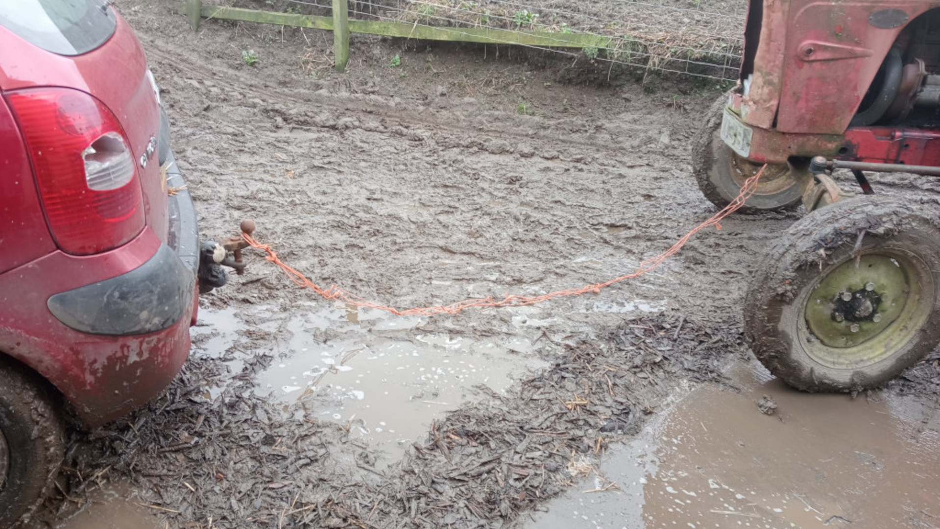 A red car stuck in the mud, being pulled out by an old red tractor using an orange rope. The scene is set in a muddy farmyard or field, with a wooden fence visible in the background. The mud is deep and thick, indicating a wet and difficult terrain.