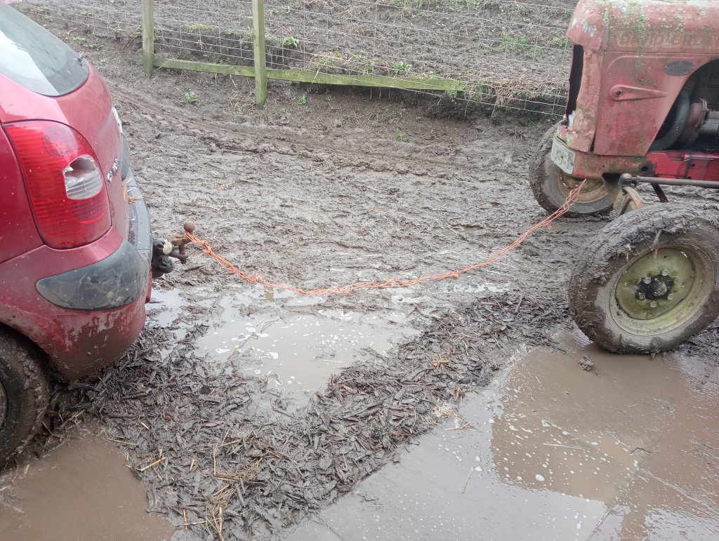 Red car stuck in the mud, being pulled out by an old red tractor using an orange rope. The scene is set in a muddy farmyard or field, with a wooden fence visible in the background. The mud is deep and thick, indicating a wet and difficult terrain.