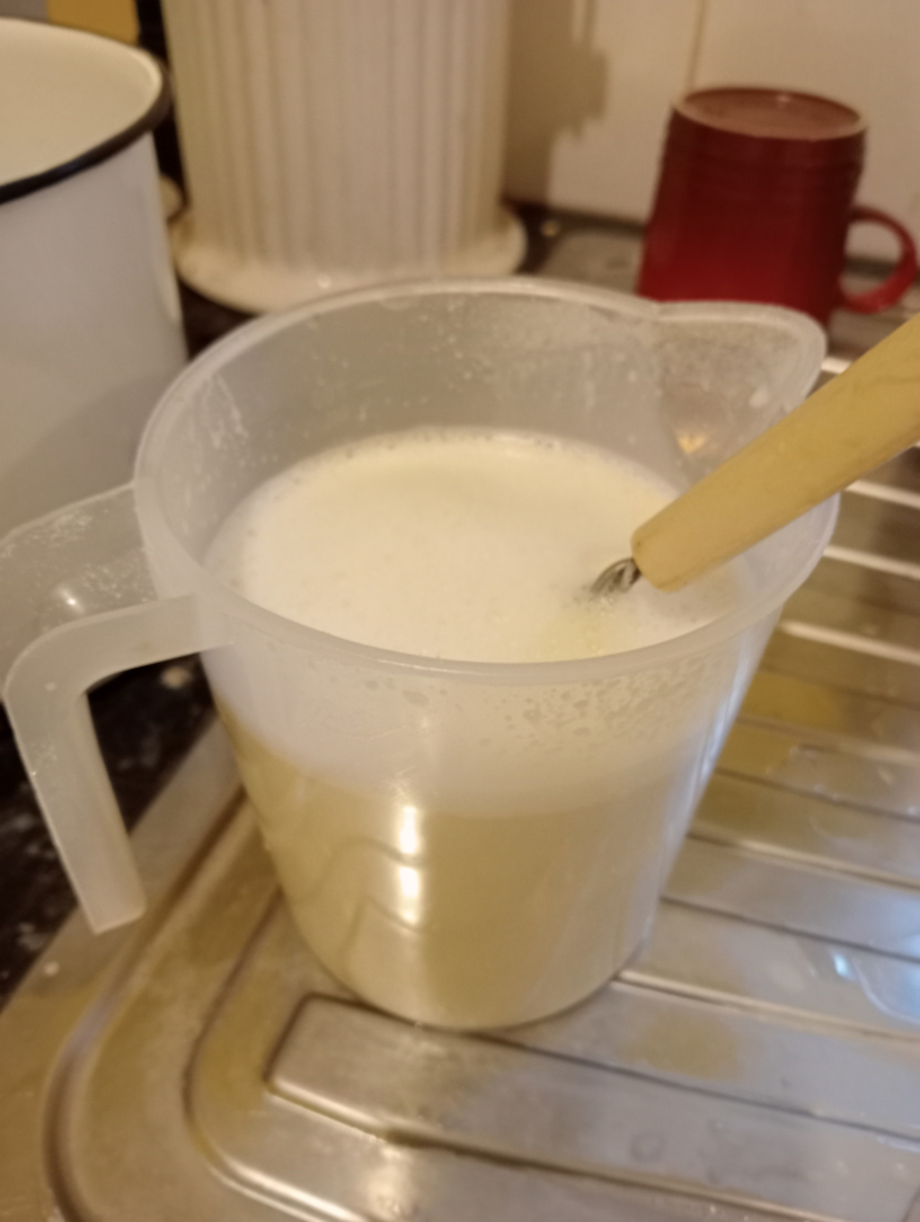 Clear plastic jug filled with a creamy, off-white liquid. A wooden whisk is partially submerged in the liquid. The jug sits on a metallic draining board in a kitchen setting, with parts of other kitchen utensils visible in the background.