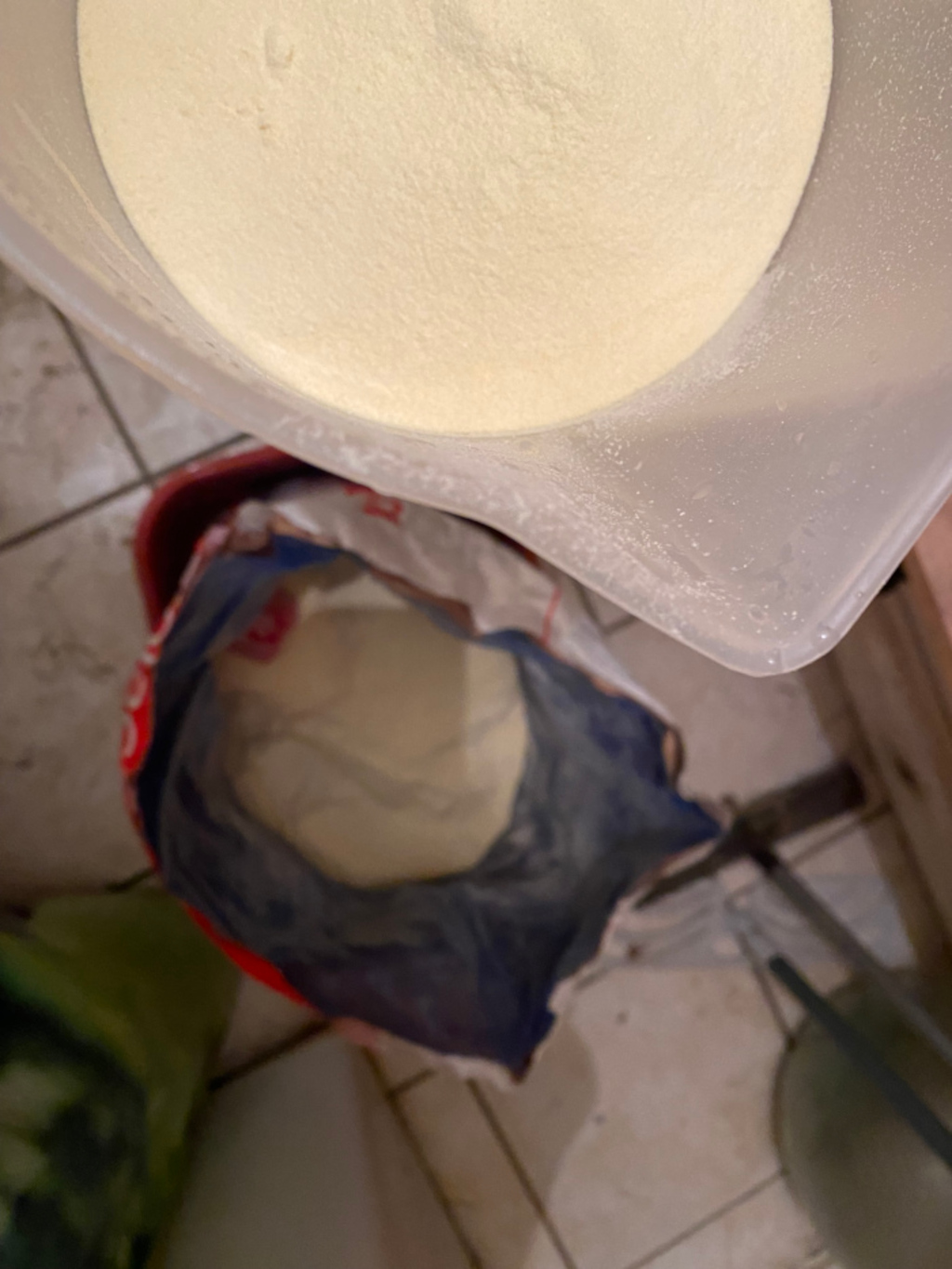 Large, clear plastic container filled with a substantial amount of a light beige, powdery substance. A portion of this substance appears to have been transferred to a partially visible, smaller blue and red bag sitting in a red bucket. The setting appears to be a kitchen or similar domestic space, evidenced by the tiled floor. The overall impression is of a food ingredient, possibly flour or a similar baking component, being measured or transferred.
