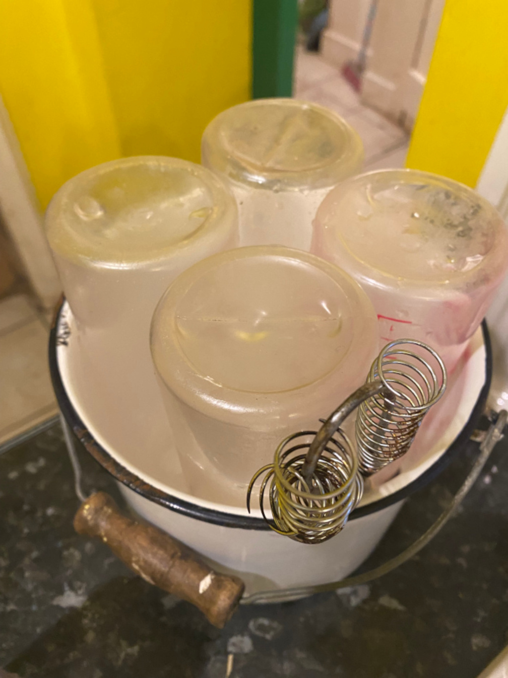 White enamel pail containing four clear glass jars. A metal whisk with a burnt appearance rests inside the pail. The pail has a wooden handle. The overall setting appears to be a kitchen or domestic environment.