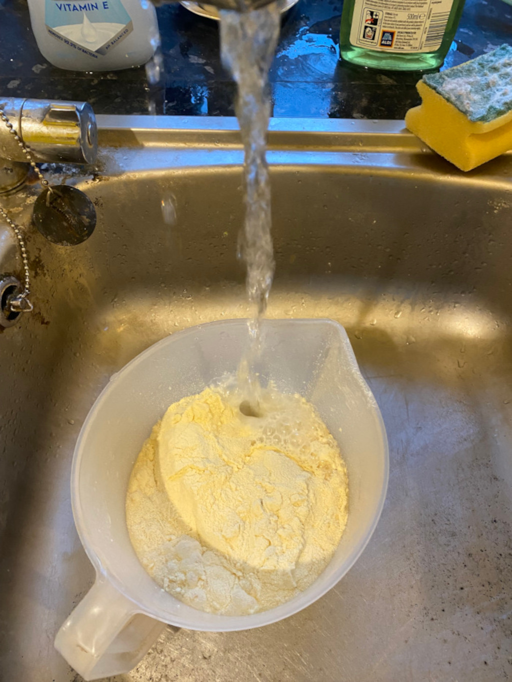 White plastic jug being filled with water from a kitchen tap. Inside the jug is a pale yellow powder, possibly flour or polenta. The sink is stainless steel and somewhat dirty. Part of a bottle of washing up liquid and a yellow sponge are visible in the background.
