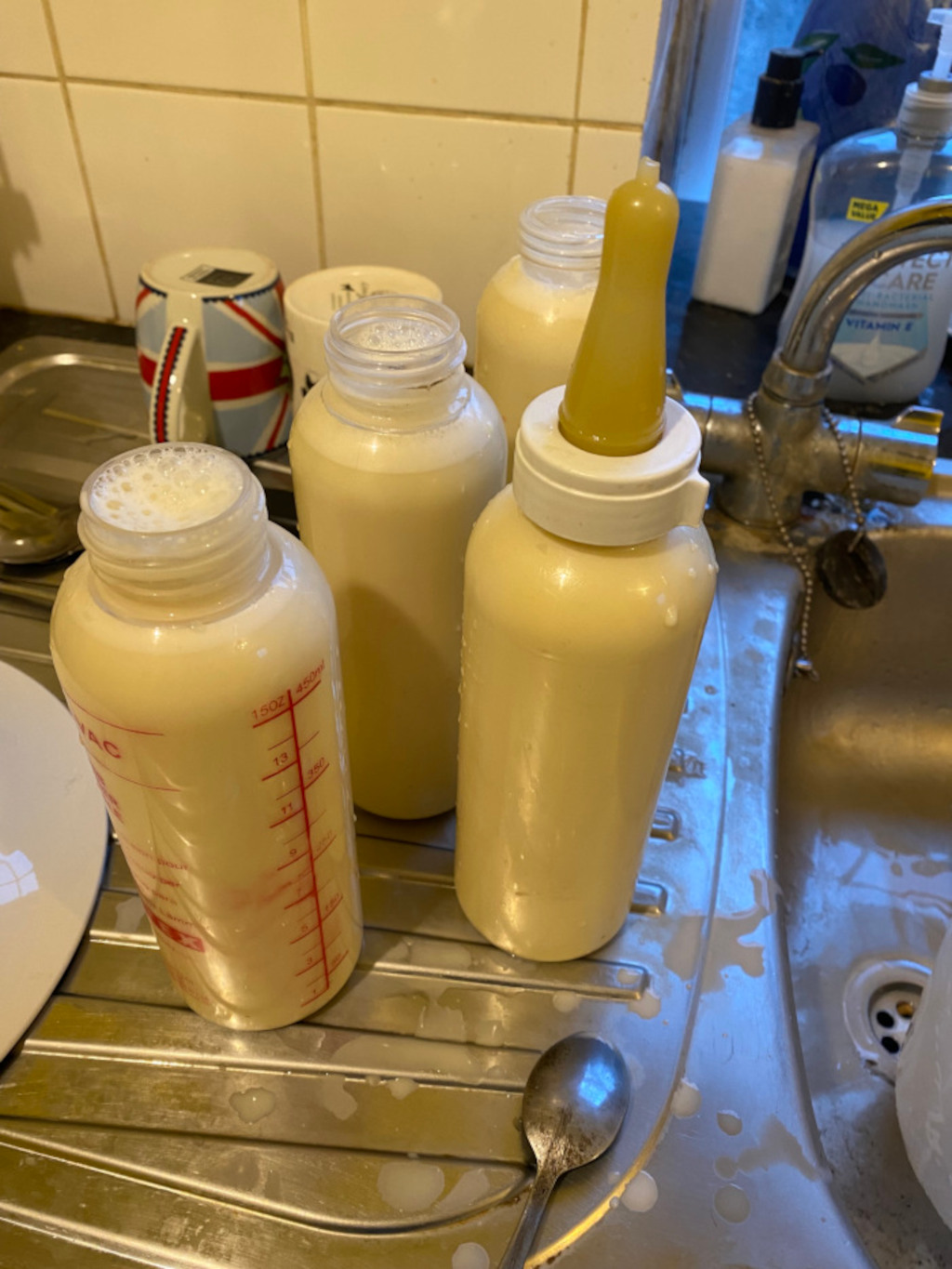 Four baby bottles filled with breast milk sitting in a kitchen sink. There are also a couple of mugs and some cleaning supplies visible in the background. Spilled milk is evident on the sink's surface.