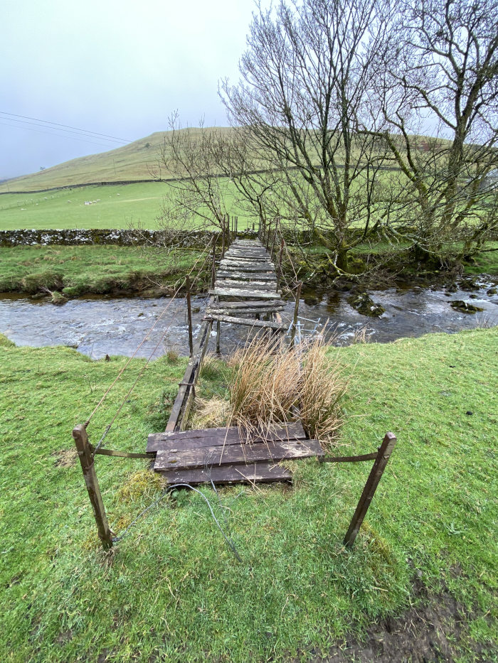 Dilapidated wooden footbridge spanning a narrow stream. The bridge is partially collapsed, with several planks missing or broken. The surrounding landscape is pastoral, with green fields, a low hill in the background, and bare winter trees along the stream bank. 