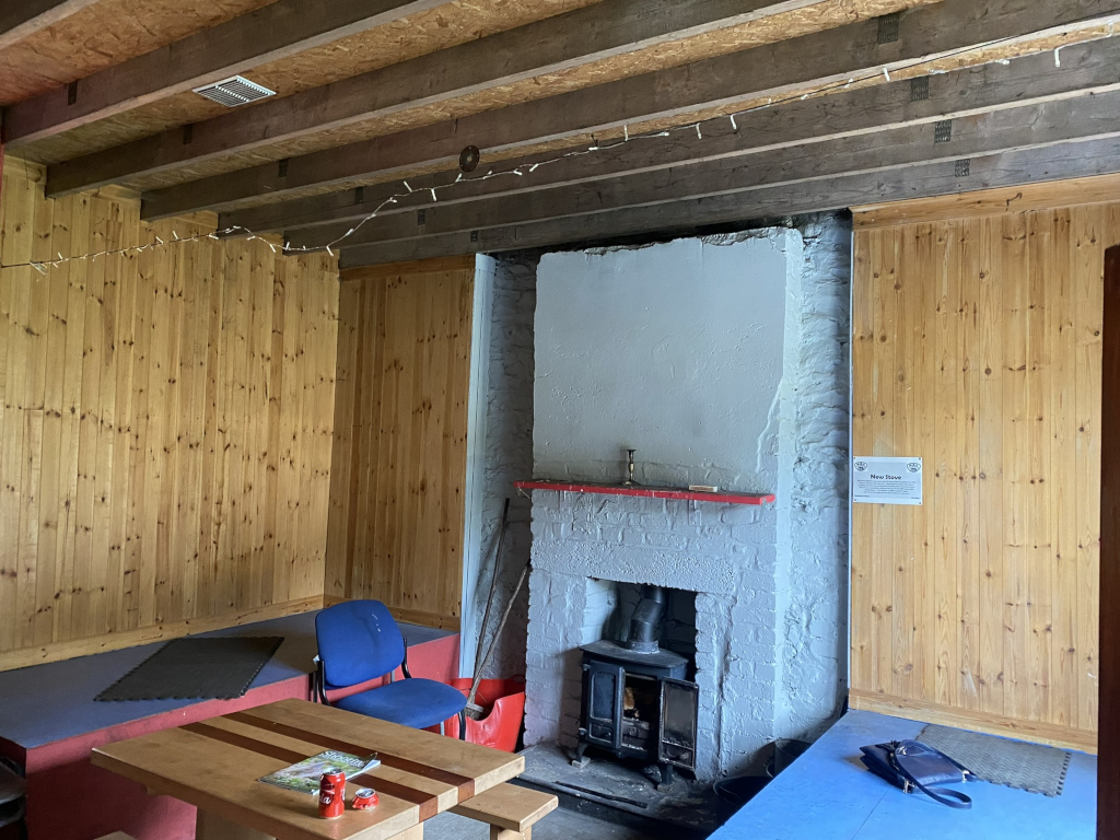 Interior of a simple, rustic room with wooden walls and a wood-burning stove as the focal point. The room appears to be a common area or possibly a small shelter, with seating provided along the walls. A simple table sits in the centre, and there are a few personal items like a purse and a magazine. String lights hang from the ceiling, adding a touch of warmth. 