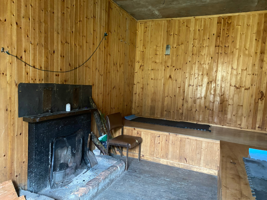 Interior of a small, rustic wooden room. The walls are panelled with light-coloured wood. A simple fireplace with a metal fireback is the room's focal point. A small chair and a low, built-in sleeping platform are present. The overall impression is one of a basic, possibly temporary shelter or a very simple dwelling.