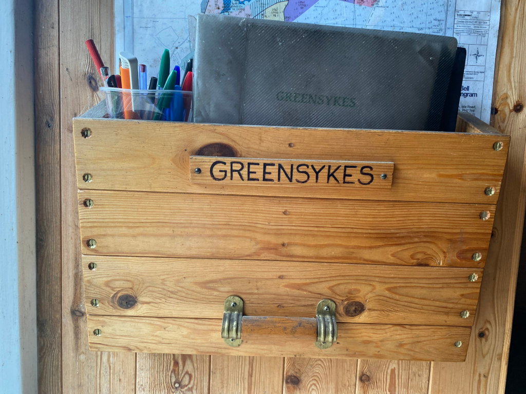 Wooden crate mounted on a wall. The crate is labelled GREENSYKES and contains various pens and pencils in a cup, along with a dark-coloured folder or file also marked GREENSYKES. Behind the crate, a portion of a map or chart is visible.