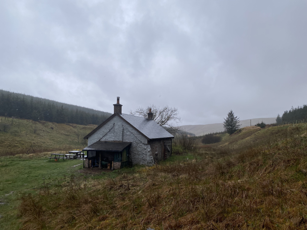 Small, stone cottage nestled in a valley, surrounded by rolling hills and sparse vegetation. The sky is overcast and grey, suggesting a cool or rainy day. A couple of picnic tables are visible near the cottage. The overall mood is serene and somewhat solitary.