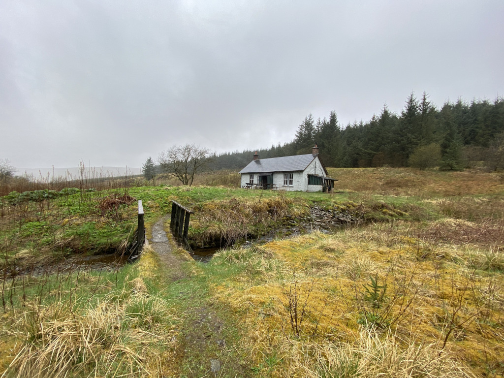 Small, white cottage nestled in a tranquil, rural landscape. A small wooden footbridge crosses a stream in the foreground, leading towards the cottage. The background features a dense evergreen forest under a cloudy sky. The overall feeling is one of serenity and isolation.