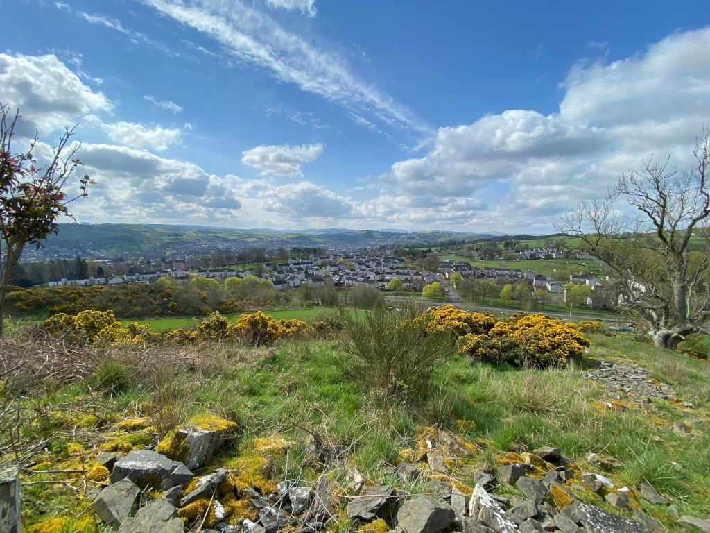 Panoramic view of a town nestled in a valley, seen from a slightly elevated vantage point. The foreground is characterised by grassy hills speckled with yellow flowering gorse bushes and scattered rocks, possibly remnants of a stone wall or structure. A leafless tree stands prominently to the right. The town in the mid-ground is composed of numerous houses and buildings, appearing relatively compact. Beyond the town, rolling hills stretch into the distance under a partly cloudy, bright blue sky. The overall impression is one of a pleasant, sunny day in a rural or semi-rural setting.
