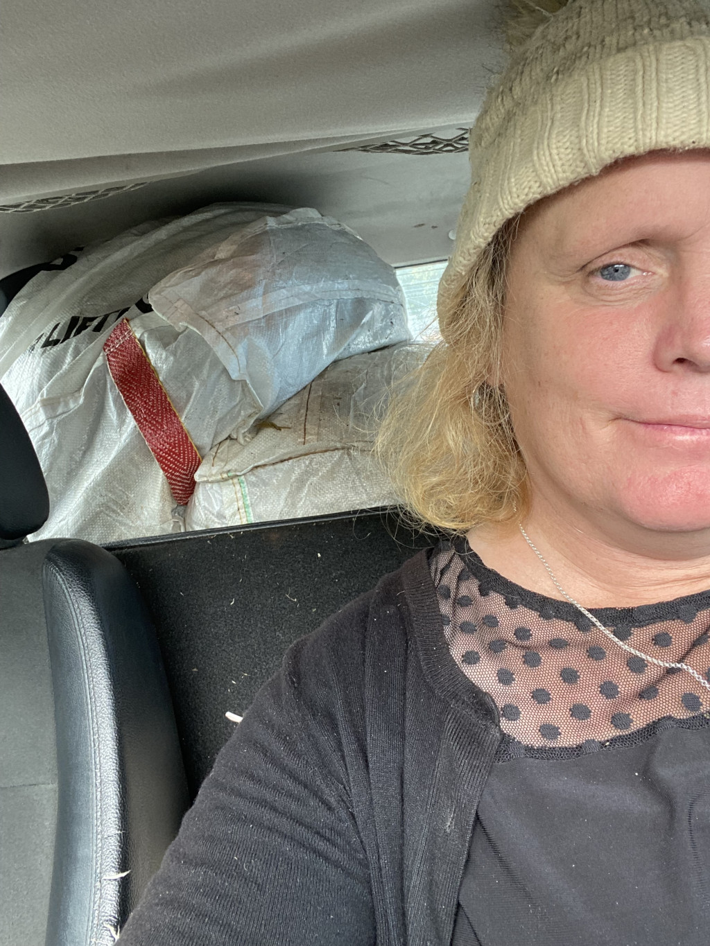 Selfie taken by a Leonie in a car. She is wearing a beige knitted hat and a black top with polka dot mesh inset. Behind her, in the back seat, are several large, dirty, white burlap-type sacks, partially obscuring the view. The overall impression is that the woman is possibly transporting a load of something heavy or bulky in her vehicle.
