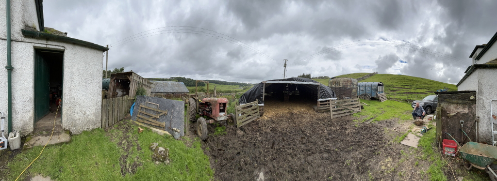 Panoramic view of a rural farm scene. In the foreground, there's a muddy area with a temporary sheep shelter covered by a black tarp.  An old tractor is partially visible near the shelter. To the left and right are stone buildings, indicative of a farm house or outbuildings. A man is seen sitting near a Land Rover parked in the yard. 