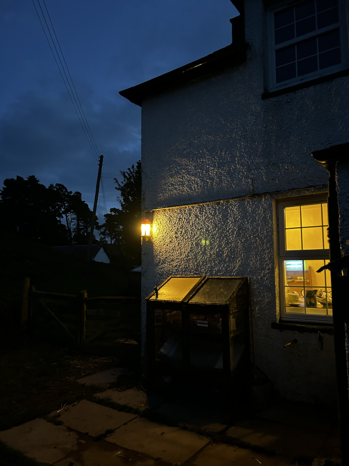 Exterior of a whitewashed building at night, illuminated by a single, warm-toned light fixture mounted on the wall. A dark, possibly wooden, structure resembling a small greenhouse or storage unit is situated against the wall near a window that shows the warm glow of an interior light. The scene is quiet and peaceful, with a dark, natural backdrop of trees and a fence suggesting a rural setting. The overall mood is one of calmness and solitude.