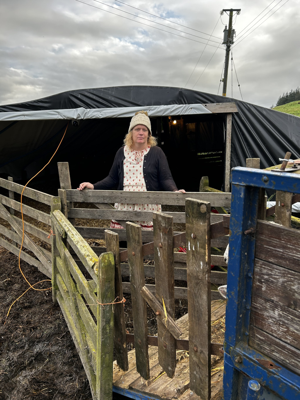 Leonie standing in front of a rustic wooden fence and gate. She is wearing a white knit hat and a polka dot dress under a cardigan. She appears to be in a farm setting, next to a simple, dark-coloured tarp structure that likely serves as a shelter or barn. A blue trailer is partially visible in the foreground to the right. The overall mood is somewhat sombre or pensive.