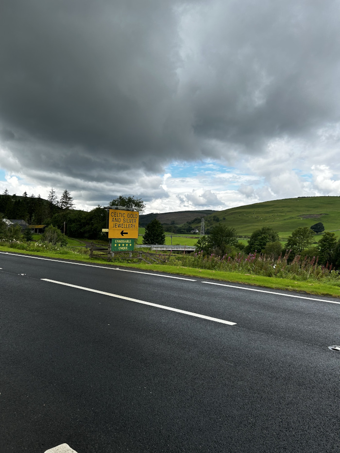Roadside view of a highway with a sign indicating Celtic Gold and Silver Jewellery and an arrow pointing to a side road. The sky is overcast with dark clouds, and the background displays a tranquil green landscape with gently rolling hills. The overall feeling is one of a peaceful rural setting, with the sign hinting at a local business or attraction nearby.