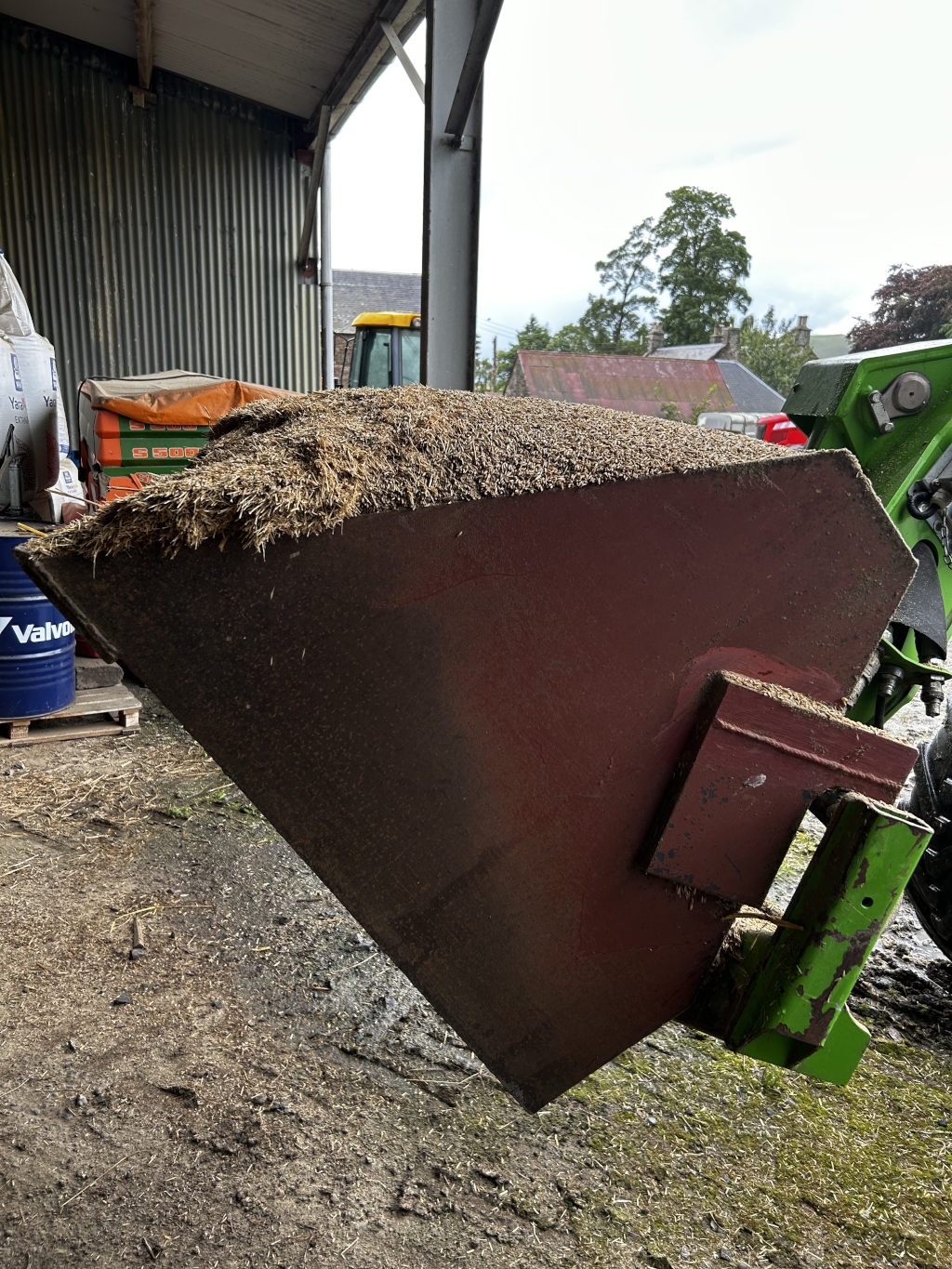 Close-up view of a partially filled metal agricultural bucket or scoop attached to a green piece of farm machinery. The bucket is filled with dried, light brown, fibrous material, presumably hay or straw. The setting appears to be a barn or farm outbuilding, with glimpses of other farm equipment and buildings in the background. 