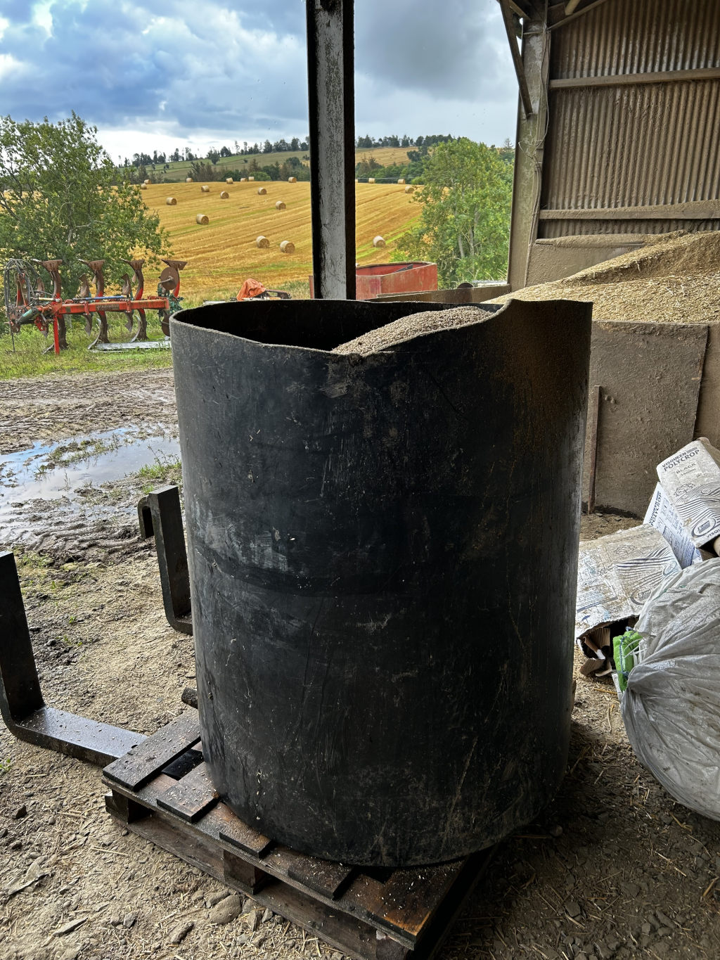 Large, black cylindrical container filled with light-coloured granular material, possibly feed or grain, sitting on a wooden pallet inside a barn. The background features a view of a harvested field with hay bales under a cloudy sky. A piece of farm equipment is visible in the distance. 