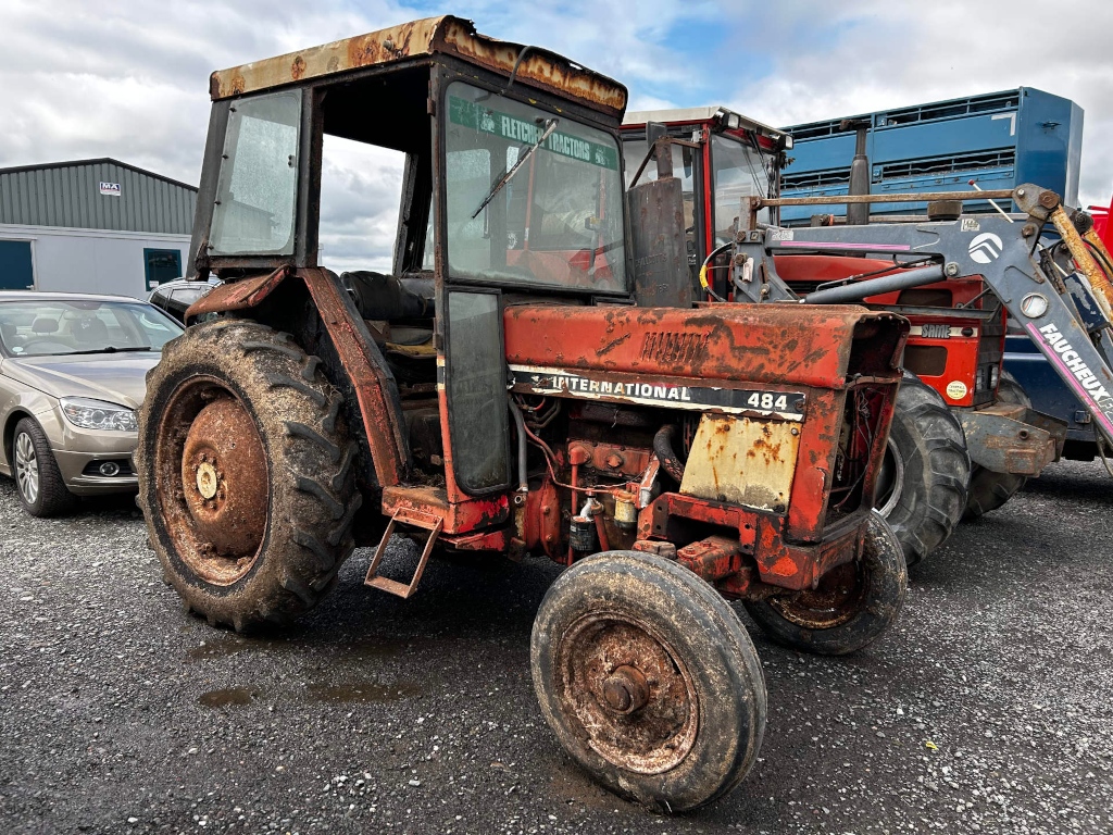 Rusty, old International Harvester 484 tractor. It's parked in a lot, near another tractor and a car, suggesting a farm or agricultural setting. The overall condition of the tractor indicates it's been used extensively and hasn't been well-maintained. The background includes a building and some other equipment, further emphasising a working, possibly rural, auction.