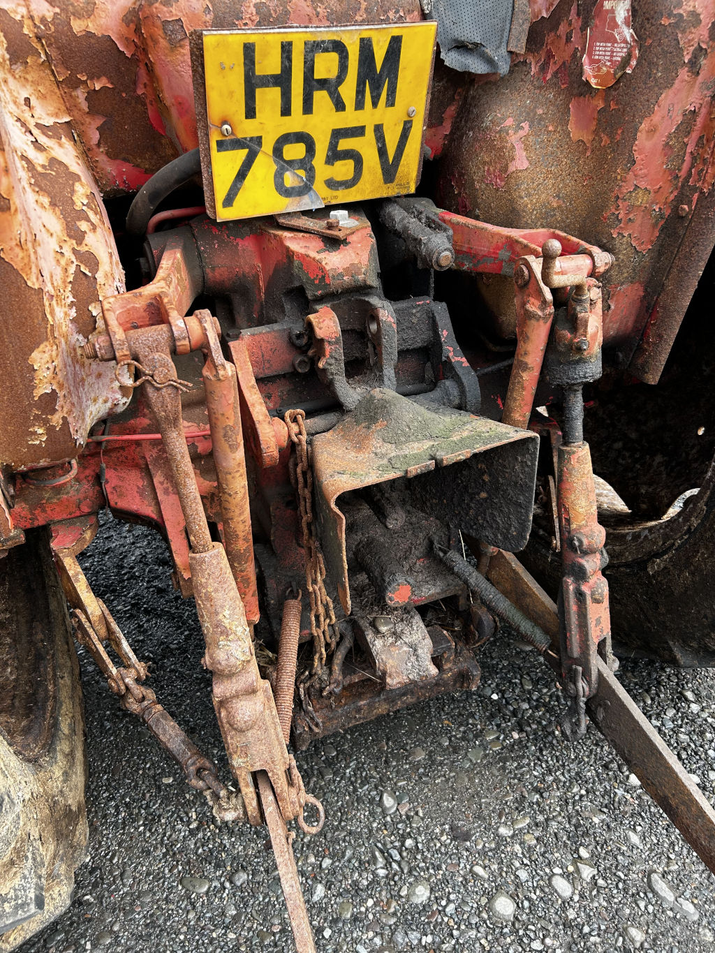 Close-up view of the rusty, heavily weathered rear hitch mechanism of an old tractor. The tractor's registration plate, HRM 785V, is visible. The overall impression is one of age, wear, and neglect, but also hints at the machine's past agricultural use.